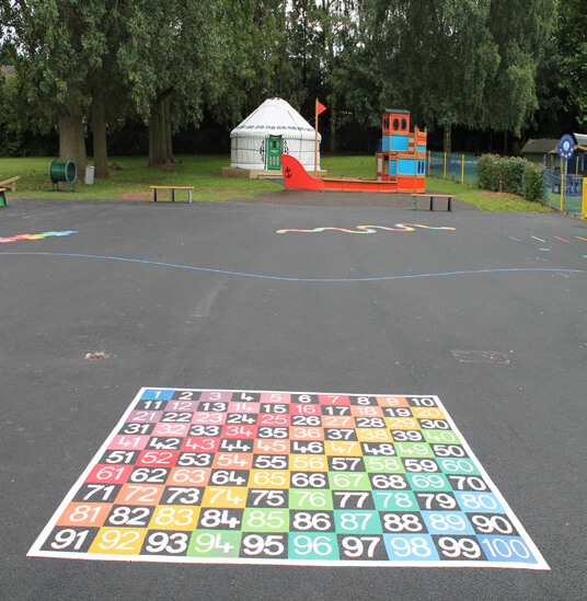 Playground at Holy Family RC Primary School with white yurt outdoor classroom, wooden boat play structure, and rainbow number counting snake and rainbow 10x10 number grid playground markings